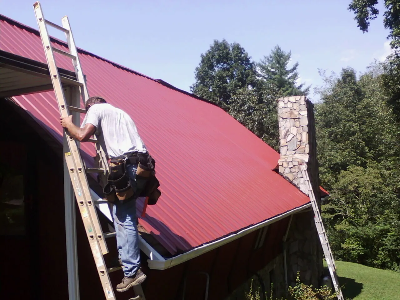Roofer on a ladder beside a red metal roof and stone chimney — Soffit & Fascia Repair by Roofing Brothers in White Plains